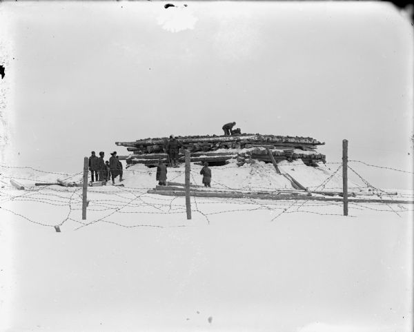 View across snow and barbed wire fence of a group of soldiers constructing a log blockhouse.
