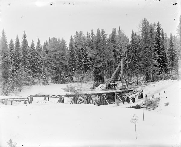 View across snow of a group of men working to construct a wooden bridge that crosses a ravine. Another group of both men and women standing on a road look on at the construction. There is a tree-covered hill in the background.