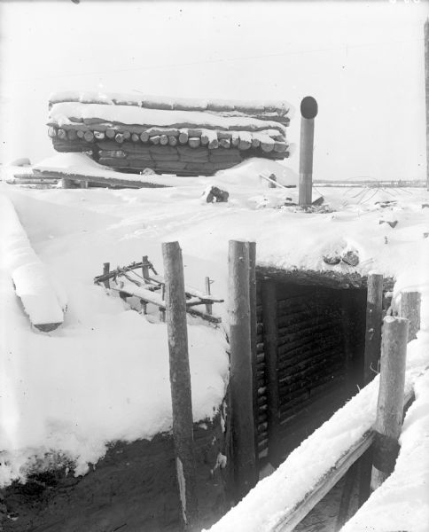 View of snow-covered blockhouse constructed of logs and sandbags, and part of a trench leading to a dugout. A stove pipe projects out of the snow above the dugout near the blockhouse.