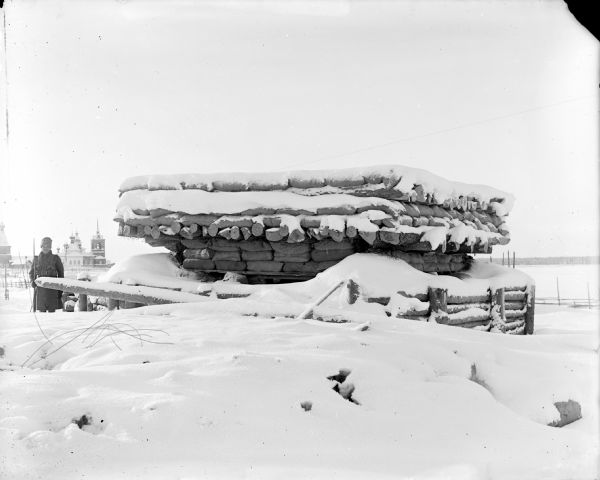 View across snow of a soldier with a rifle standing near a timber and sandbag constructed blockhouse. In the distance is a Russian town. The most prominent structures appear to be churches with onion domes.