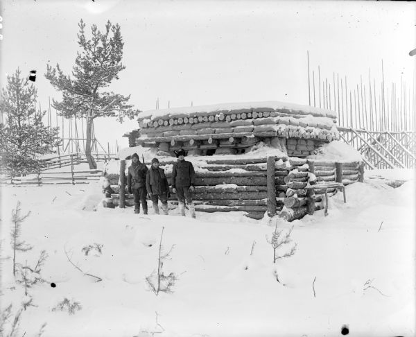 View across snow of three soldiers standing in front of a blockhouse that has a tall wooden fence along the exterior. The man on the left is armed with a rifle and the soldier on the right is wearing an American uniform.