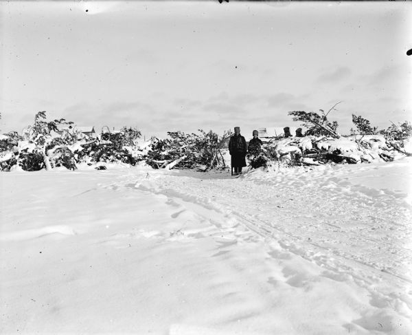 View across snow towards four soldiers standing near brush piles. Two men standing in the center, and two men stand behind the brush, defending the road to the front. There are wooden buildings in the background behind the group.