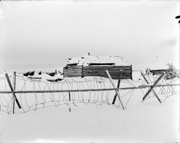 Two timber constructed blockhouses in the snow behind a barbed wire fence.
