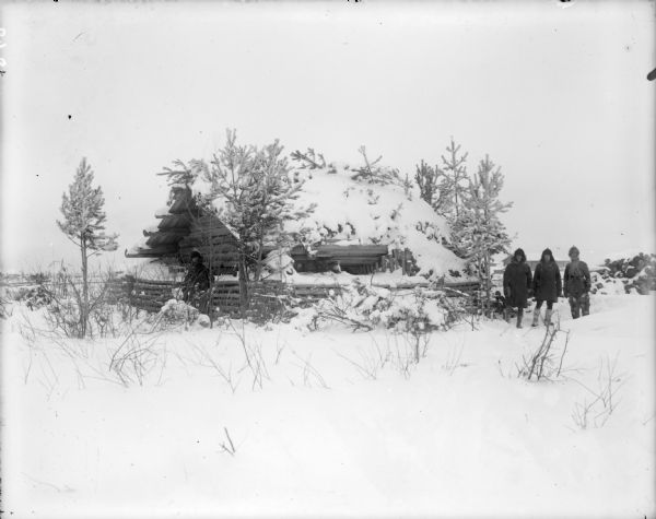 Four soldiers stand outside a blockhouse in the snow. One man stands on the left side of the building, the other three men stand on the right. The building is partially concealed by small trees and snow-covered branches piled on the roof. There are other buildings in the far background.