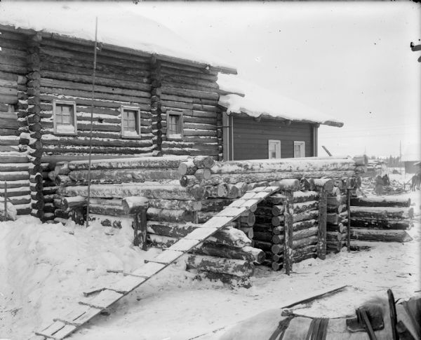 A shell-proof shelter alongside other buildings, possible a soldiers' barracks. In the distance there are two men placing wood on a horse-drawn sled. Another horse is in the foreground on the right.
