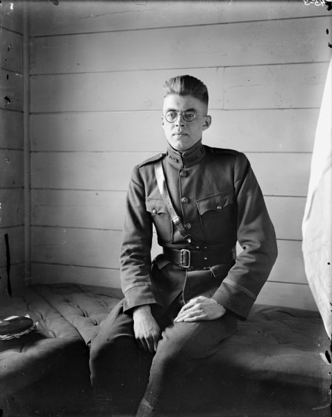 A portrait of First Lieutenant Edward W. Legier, Company C, 310th United States Army Corps of Engineers, in uniform and sitting in the barracks. A stack of phonograph records is on the cushion beside him.