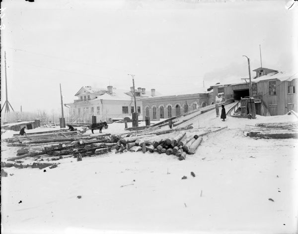 View across snow-covered ground towards a saw mill operated by the U.S. Corps of Engineers. Soldiers are loading logs to be processed on to a horse-drawn sled.