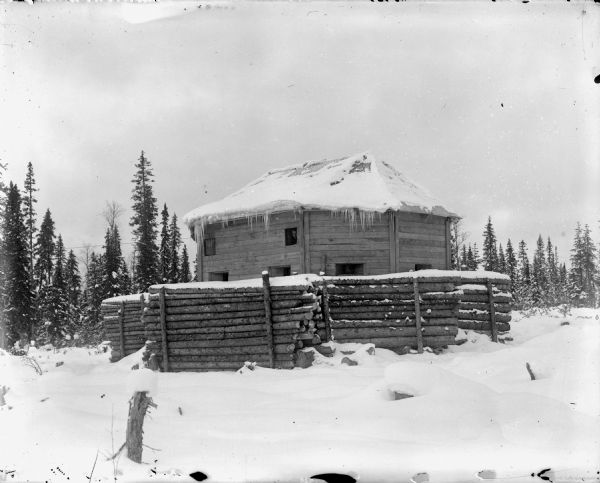 An eight-man blockhouse fortification in the snow, with a built-up wall of logs surrounding it. Soldiers inside the fortification are looking out of some of the windows. The blockhouse was used to help defend Allied soldiers against Bolshevik forces.