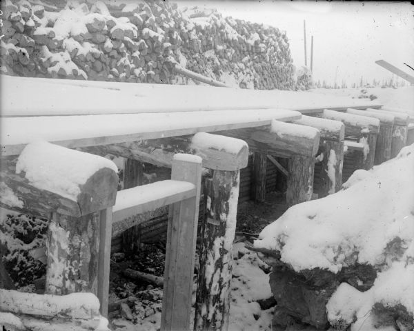 A dugout under construction, showing the timber frame and planks of wood over the top. On the left of the dugout is a large pile of timbers covered with snow.