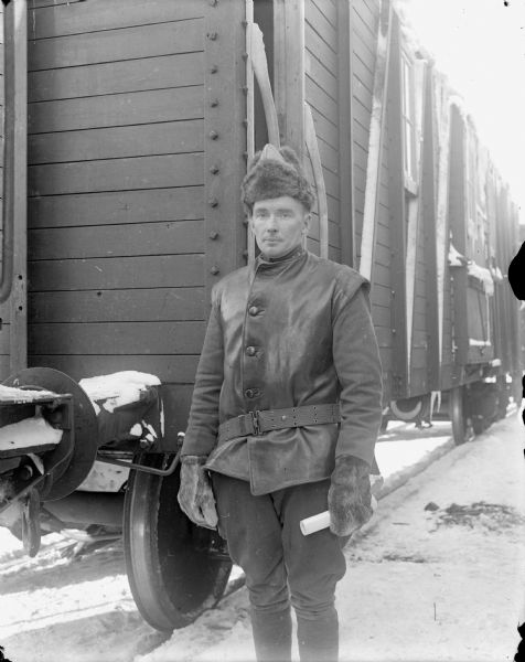 A portrait of Lieutenant W.C. Giffels of the 339th United States Army Engineer Corps, A Company. He standing in front of a railroad car and is wearing a fur hat and mittens, and a leather vest over his jacket.