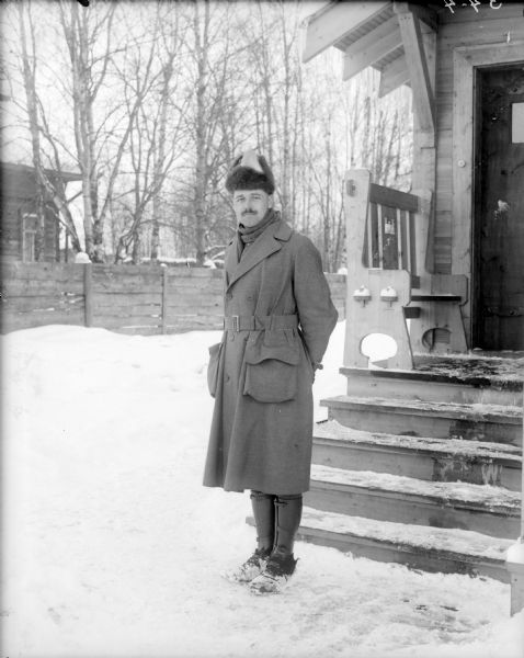 Outdoor portrait of Lieutenant R. Jens of Company C, 310th United States Engineers. He is wearing an officers great coat, leggings, and a fur hat. Behind him on the right is a wood building with a small front porch with a bench which has the United States Army Engineers emblem carved out of the back. In the background across the snowy yard is a wooden fence and behind it another building.
