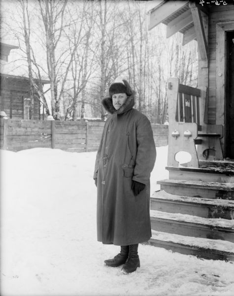 Outdoor portrait of Lieutenant H. Pryale of the 310th United States Army Engineer Corps, C Company, wearing a heavy coat and gloves, as well as a fur hat. Behind him on the right is a wood building with a small front porch with a bench which has the United States Army Engineers emblem carved out of the back. In the background across the snowy yard is a wooden fence and behind it another building.