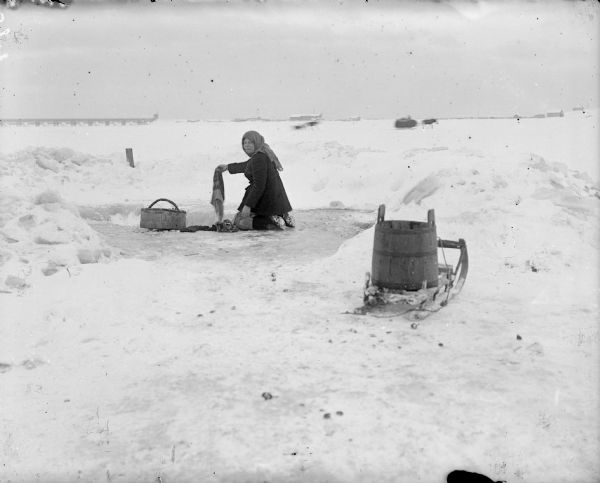 A Russian woman wearing a head scarf is doing laundry while kneeling on the frozen Northern Dvina River, near Arkhangelsk, Russia. It appears she used a small sled to carry the laundry and two large wooden pails to the river. In the distance there are two sleds being pulled by a beast of burden. There is also a bridge that crosses the river and a settlement on the opposite shoreline.