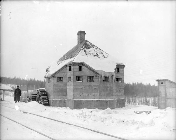 A large concrete blockhouse fortification at Verst 581 with  a concrete outhouse next to it on the right. An armed solder wearing winter clothing stands between the fortification and railroad tracks.  This fortification was constructed when the 310th United States Army Engineer Corps was deployed as a part of the Allied Intervention in Northern Russia.