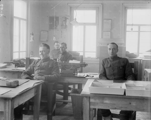 A group portrait of four soldiers in uniform sitting at their desks at the 310th United States Army Engineer Corps headquarters office. The soldier on the far back on the left side has the stripes on his uniform of a corporal. The two framed portraits next to the window appear to be General John J. Pershing and President Woodrow Wilson. The large typewriter is an Underwood Standard Typewriter.