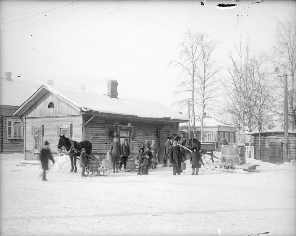 View across snow towards a group of Russian men, women, children, and American soldiers at a water station. Behind the sled on the left is a dog, and horses are pulling sleds used to carry large wooden barrels of water. The water station is surrounded by other houses or buildings.