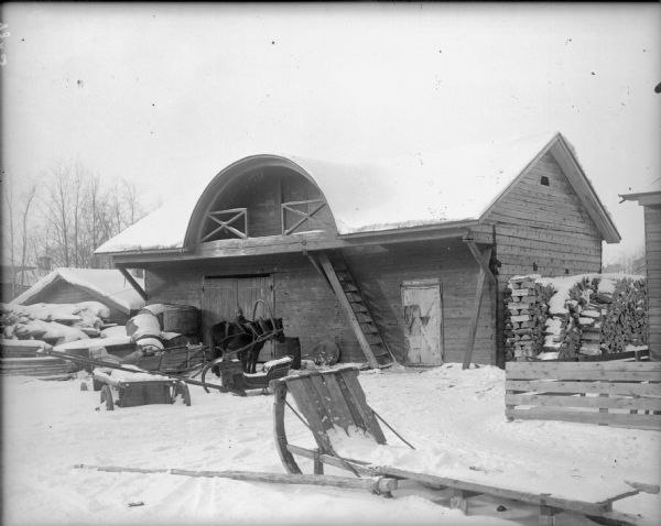 A Russian man is standing with his horse in front of a large barn. The barn has an arched balcony above the double doors. Around the barn half-buried in the snow are horse-drawn carts, sleds, and large wooden barrels.