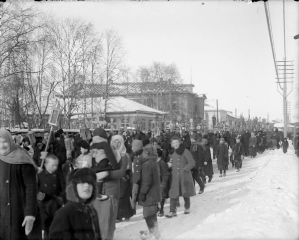 View down side of street of a large group of Russians conducting a religious parade through a town. Many of the participants carry large crucifixes and banners.
	