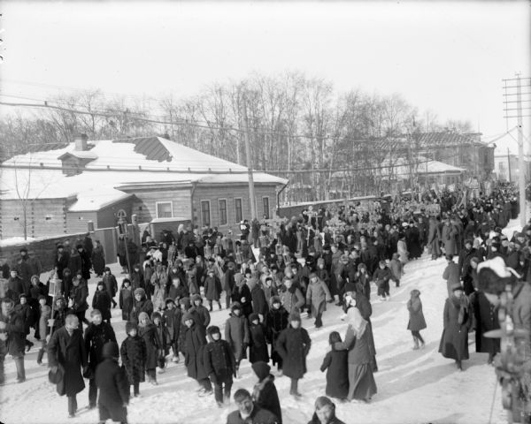 Elevated view of a large group of Russians conducting a religious parade through a town. The men in the group are carrying the iconography. Soldiers in military uniforms are near the wooden fence on the left.