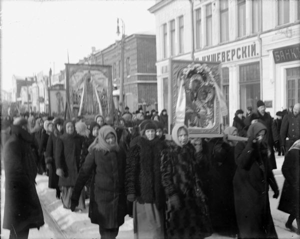 A large group of Russians conducting a religious parade through a town. The women in the group are carrying the iconography on their shoulders.