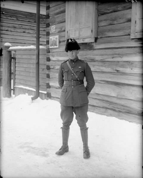 Outdoor full-length portrait of Captain McArdle from Company A,       of the 310th United States Army Engineer Corps. He is standing in the snow in front of the Headquarters of the 310th Army Engineer Corps. The log building behind him has wooden shutters which are closed, and a sign posted which says: "HQ. 310".