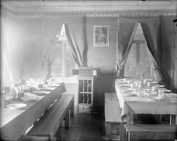 The dining room for the headquarters detachment of the 310th U.S. Army Engineer Corps while it was deployed to Archangel (Arkhangelsk), Russia. A portrait of General John Pershing hangs on the wall between two windows above a with a record player. Two tables are laid with flatware and china.