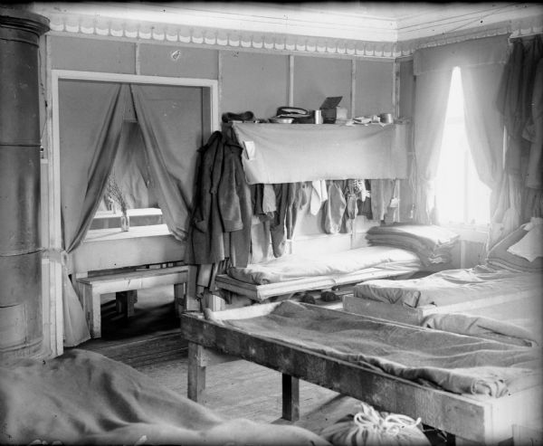 The interior sleeping quarters for the headquarters detachment of the 310th U.S. Army Engineer Corps. In the room are sleeping bunks, a woodstove, and clothes hanging from a shelving unit. The benches and tables of the dining room can be seen through a curtained doorway.