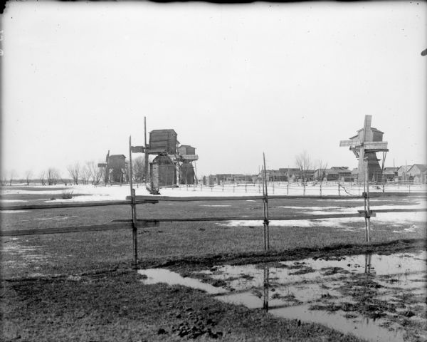 Four large wooden windmills along a wooden fence. Behind the windmills are the buildings of a village.