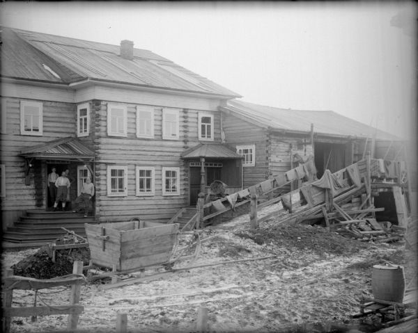 Exterior view across yard of a two-story house with a barn built adjacent to it. Two people are standing near the door of the house. In the yard is a wooden sled, a barrel, and clothes hanging out to dry along the hand railings to the barn.