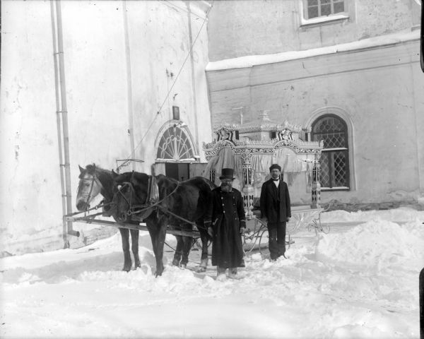 Outdoor portrait in front of a building of two men standing in the snow with a decoratively carved winter hearse complete with curtains. The driver and the assistant are standing alongside the hearse which has a team of two horses.