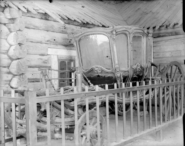 The carriage used by Peter the Great is stored alongside a log house, within a fenced-in area.