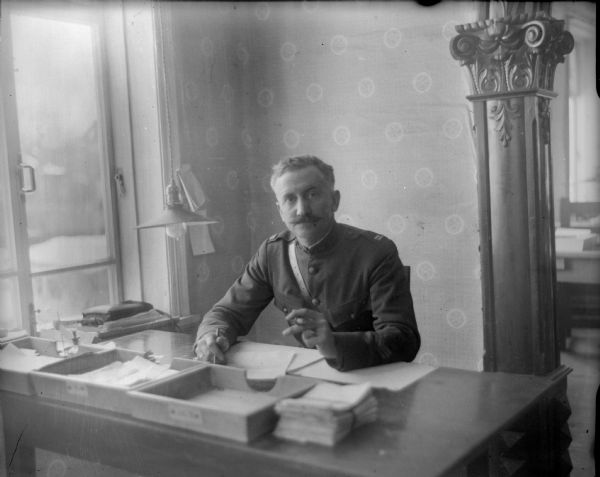 Seated portrait of Captain B.D. Gutherie, the supply officer for the 1st Battalion, 310th United States Engineer Corps. He is wearing his uniform and is sitting at his desk holding a pen in one hand and a cigar in the other. 
