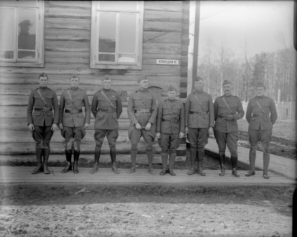 Group portrait of the headquarters staff officers of the 310th United States Army Engineer Corps. Included in this portrait are Captain McArdle of Company C (third from the left) and Lieutenant Colonel P.S. Morris (fourth from the left). The eight men are standing on a board sidewalk in front of a log building.