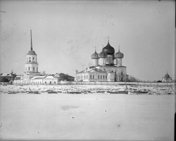 Exterior view across ice of a Russian Eastern Orthodox church with onion domes. There are a number of outbuildings as well as a fence surrounding the church.
