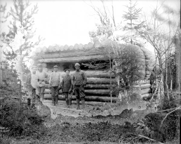A log constructed blockhouse for machine gun emplacement surrounded by trees. In front of the fortification are four men posed standing. They are possibly soldiers out of uniform, one of which appears to be wearing an Imperial Russian Army cap. Four of the men are holding axes.