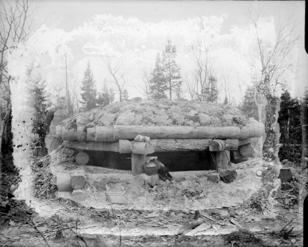 Exterior view of dugout machine gun emplacement. There is a solider standing outside on the left, while another man sits inside. The fortification appears to be primarily constructed out of logs, with the use of earth to help provide additional protection around its base and on the roof. A satchel and a large pot with a spout are sitting at the entrance to the blockhouse.
