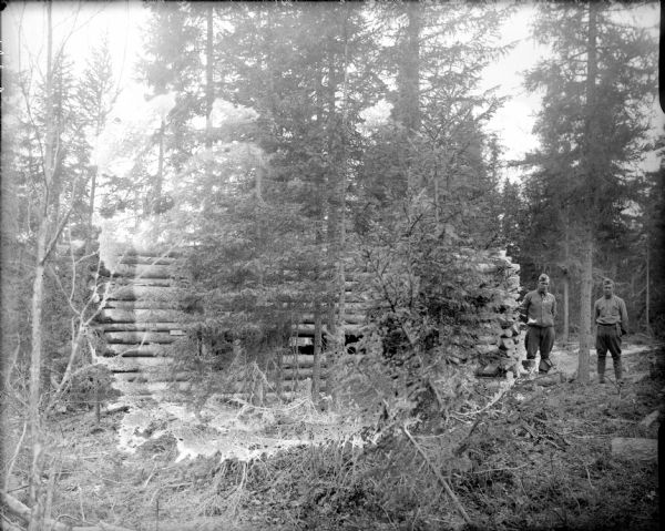 Blockhouse fortification under construction in a forested area. There are two uniformed soldiers standing on the right.