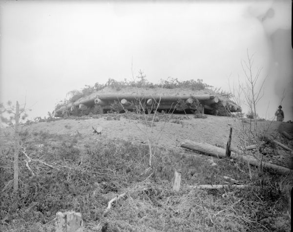 View looking up hill towards Blockhouse No. 1 that is constructed from timbers and earth. The fortification appears to be partially constructed into the top of a hill. A uniformed solider stands on the hill on the right.