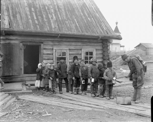 Russian children lined up on a board sidewalk to receive leftovers from a man standing outside the 310th United States Army Engineer corps mess hall. Some of the children are holding tins to receive the leftovers. In the background is a woman and child, and a dog sitting on the ground.