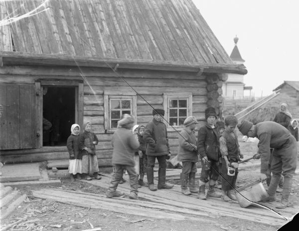 Russian children lined up on a board sidewalk to receive leftovers from a man standing outside the 310th United States Army Engineer corps mess hall. Some of the children are holding tins to receive the leftovers. In the background is a woman and child, and a dog sitting on the ground.