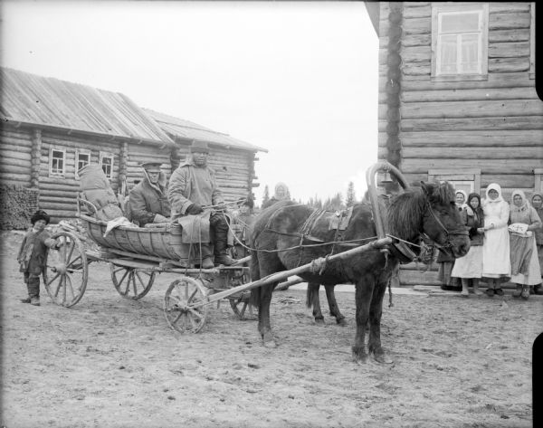 Major McArdle returning to a village from an inspection trip. He is riding in a horse-drawn wagon with a driver, and local Russians, women and children, are standing nearby. In the background are a number of log buildings.