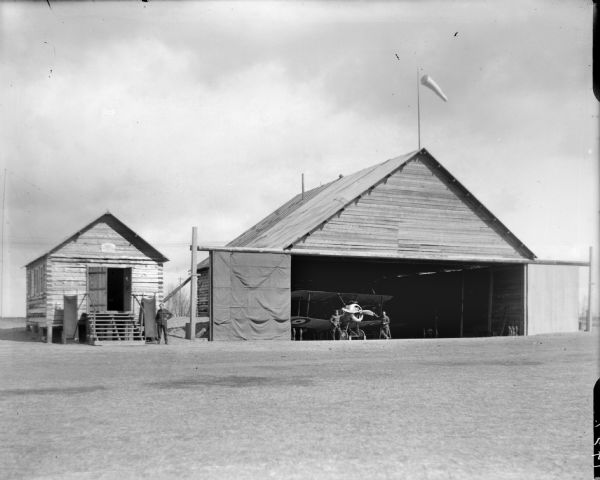 Airplane hanger constructed by Company B from the 310th United States Army Engineer Corps. One soldier is standing in front of the small building next door to the airplane hanger. Two stretchers are propped on either side of the doorway. Two soldiers are standing inside the hanger alongside a Sopwith Camel aircraft. The hanger has an open door, which appears to be made of canvas.