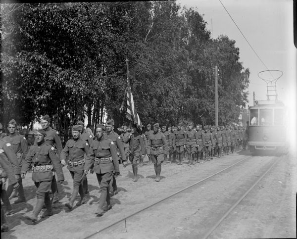 DESCRIPTION QUESTION: A group of American soldiers are marching down a street in Archangel [Archangelsk], Russia. Next to the flag barrier is a African American uniformed soldiers. Passing the soldiers on their left side is an electric street car.

ANDY--not sure about this description. Did Chad mean to say "flag bearer"? I see one man next to the person carrying the flag, and I would not be sure he is African American.