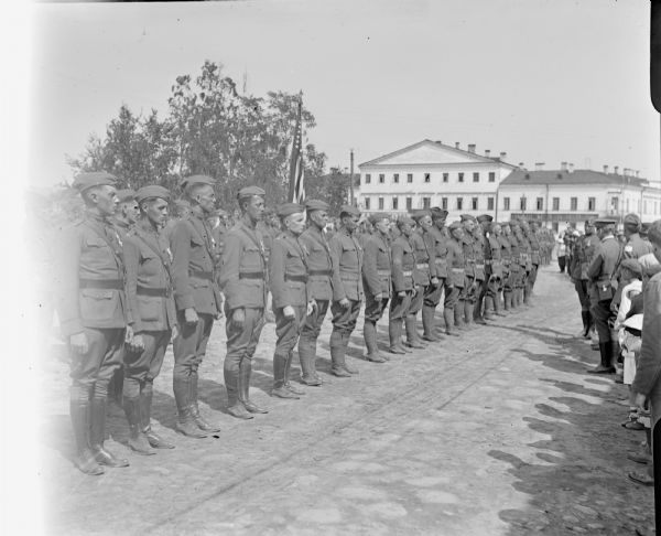 A group of uniformed soldiers and officers from the 310th United States Army Engineers standing at attention for review by commanding officers. Amongst the group of men at attention is Major McArdle, standing on the far left. On the right side is a group of other officers and civilians.