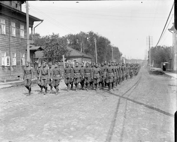 View of an approaching group of soldiers coming up a road. A captain flanked by a lieutenant on the right is leading the group of soldiers from the 310th United States Army Engineer Corps on a march through a town.