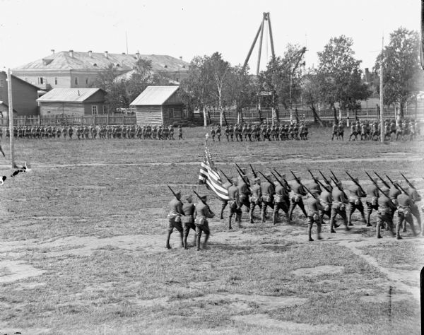 Slightly elevated view of groups of soldiers performing drills in a field surrounded by a fence. At the rear of the marching column one man carrying an American flag is flanked by two men carrying rifles. There are a number of buildings in the background, including tall crane-like structures.