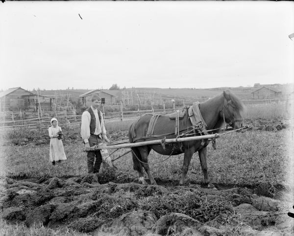 A peasant farmer plowing a field with the plow harnessed to a horse. The farmer's daughter is wearing a light-colored patterned dress and stands on the left holding a plant. In the distance are a number of log constructed buildings with wooden fencing around them.
