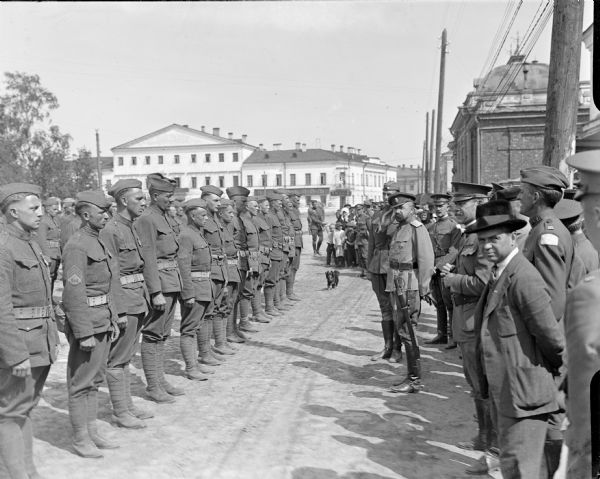 A group of soldiers from the 310th United States Army Engineer Corps standing at attention on the left for inspection of American and Russian officers in the town square. The officers reviewing the soldiers are Major General William S. Graves (third from the right), commander of the American Expeditionary Forces in Siberia, Russia, Russian General Lavr Kornilov (fourth from the right), and Lieutenant Colonel P.S. Morris (fifth from the right). A small dog stands in front of a group of children in the background.