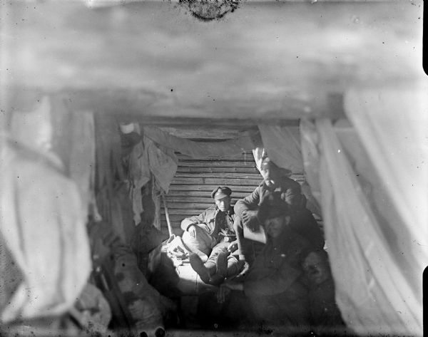 Group portrait of either British or American uniformed soldiers sitting in a dugout. Fabric, either curtains or clothing, is hanging down from the low ceiling.