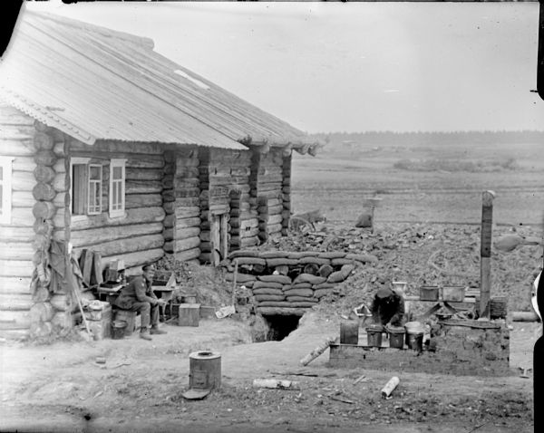 Two soldiers outside of a trench that is next to a log cabin which is reinforced with sandbags. One of the soldiers appears to be preparing a meal in an outdoor kitchen, while the other soldier sits on a log. There is a field in the background.
